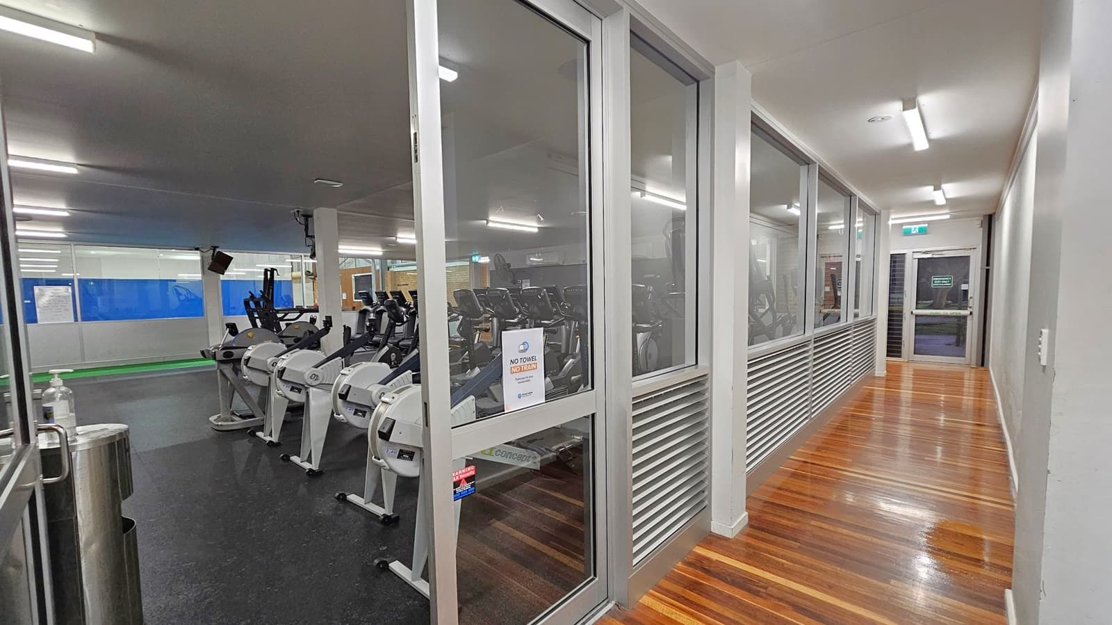 A gym interior with a row of exercise bikes on the left, visible through large glass windows. The floor is dark and rubberised. On the right, a corridor with polished wooden flooring leads to a door at the end. The ceiling is fitted with fluorescent lights, and a sign on the window reads "No Towel No Train".