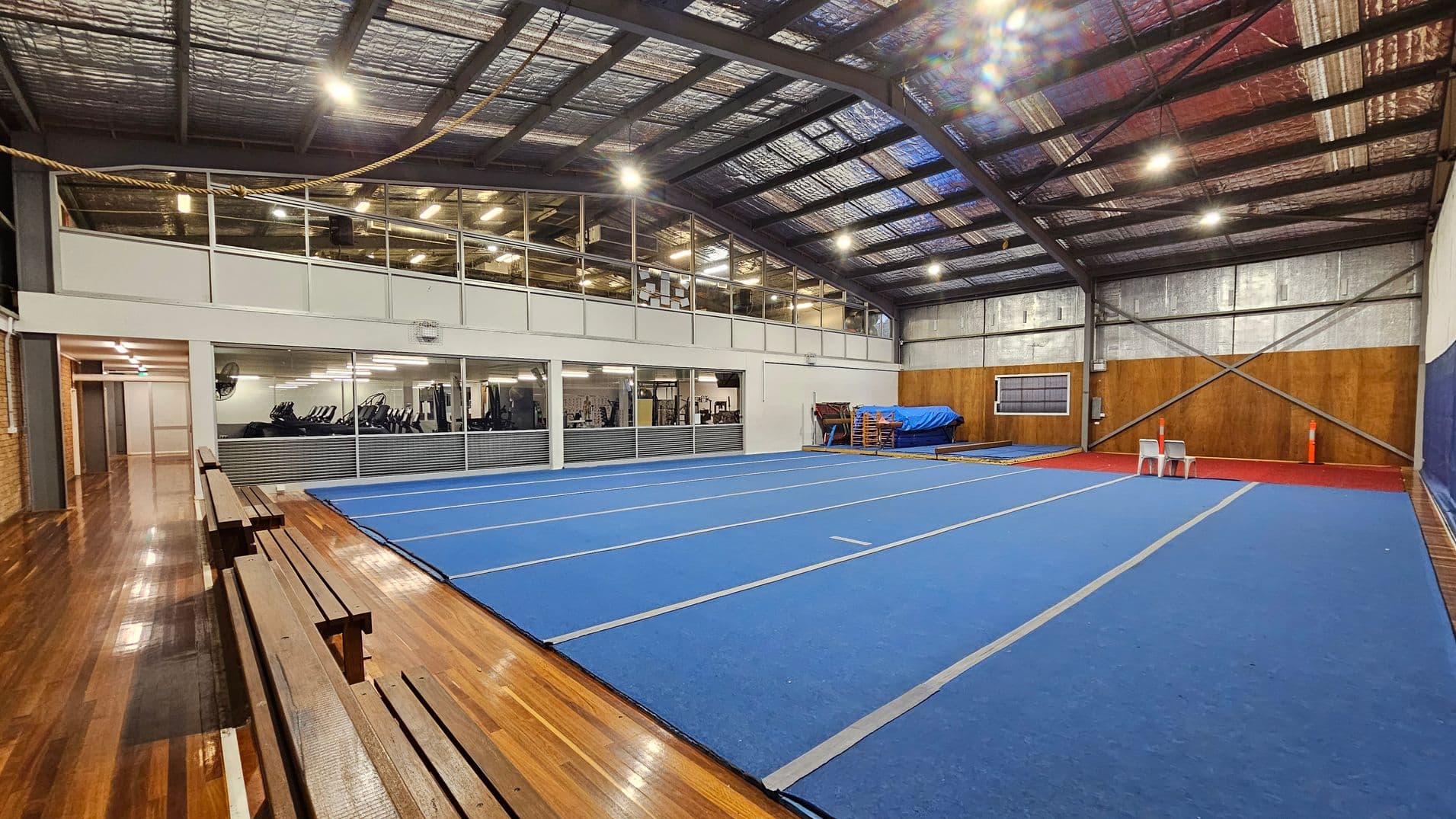 A spacious indoor gymnasium with a blue mat covering the floor in the centre. On the left, there are wooden benches along a polished wooden floor. The right side features a wall with wooden panels and two white plastic chairs. In the background, a mezzanine level with glass windows overlooks the gym, revealing exercise equipment inside. The ceiling is high with exposed metal beams and insulation.