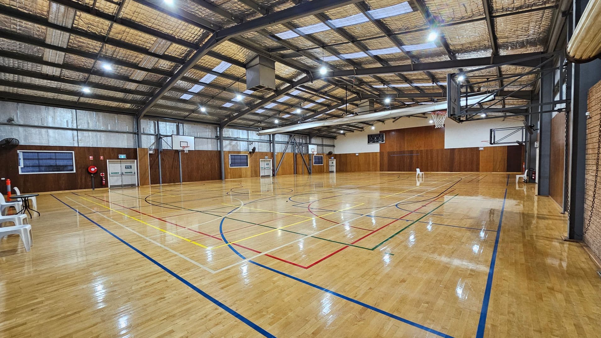 A spacious indoor basketball court with a polished wooden floor marked with various coloured lines. On the left, there are white plastic chairs and a table against a wall with a fire extinguisher and exit doors. The right side features a basketball hoop attached to the wall. The ceiling is high with exposed beams and lighting, and the walls are lined with wood panelling.