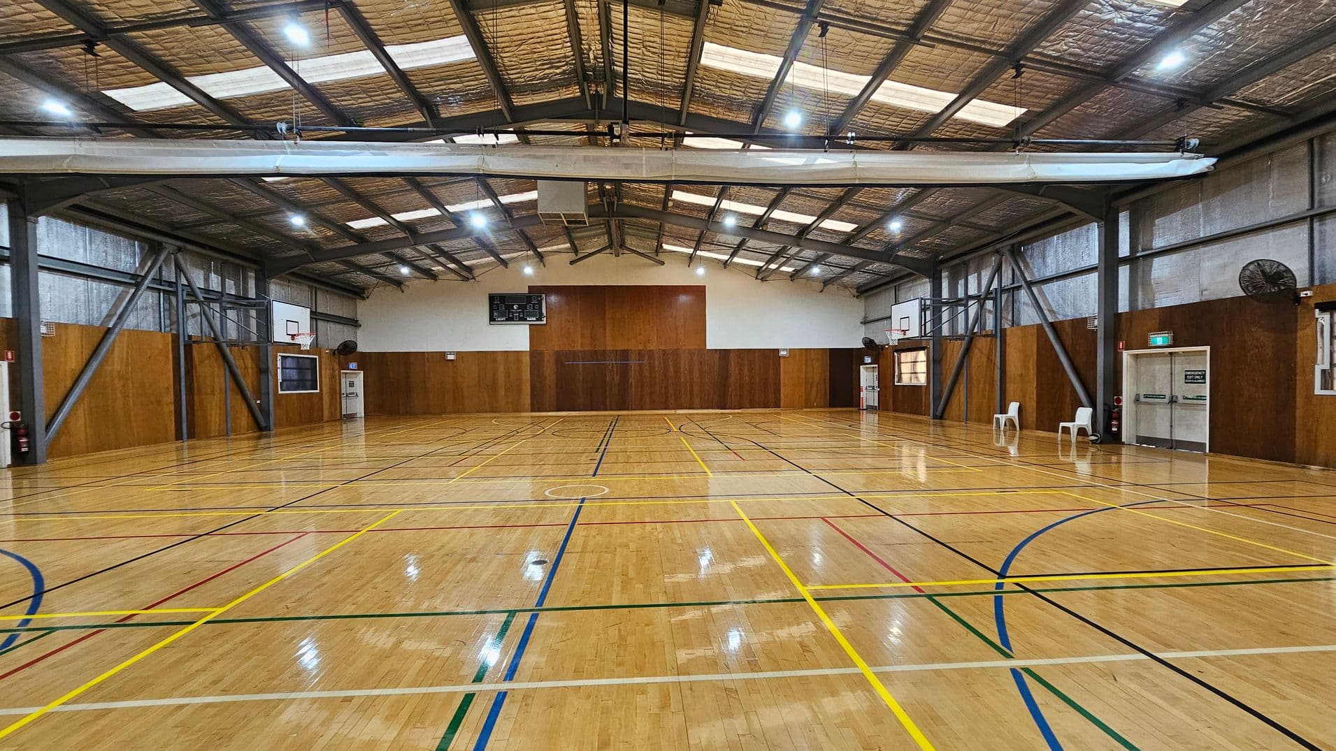 A spacious indoor basketball court with a polished wooden floor marked with various coloured lines. Two basketball hoops are mounted on the walls at each end. The walls are lined with wood panelling, and there are metal beams supporting the structure. The ceiling is high with exposed insulation and lighting. On the right, there are two white plastic chairs near a set of double doors. A scoreboard is mounted on the far wall.