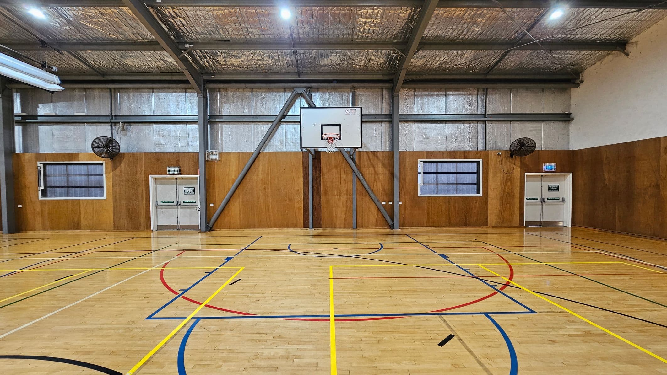 A gymnasium with a wooden floor marked with various coloured lines for sports. In the centre is a basketball hoop attached to a metal frame on a wooden-panelled wall. Two large fans are mounted on the wall, one on each side of the hoop. There are two doors with exit signs, one on the left and one on the right. The ceiling is high with exposed metal beams and insulation. Two windows are visible, one on each side of the hoop.