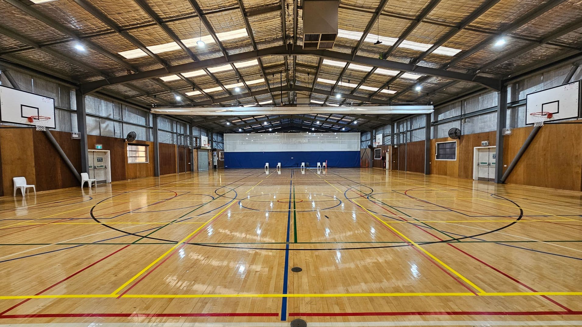 A spacious indoor basketball court with a polished wooden floor marked with various coloured lines. On the left and right, there are basketball hoops mounted on the walls. Several white plastic chairs are placed along the sides. The ceiling is high with exposed beams and lighting fixtures. The walls are a mix of wood panelling and metal, with ventilation fans visible. At the far end, a blue wall serves as a backdrop.