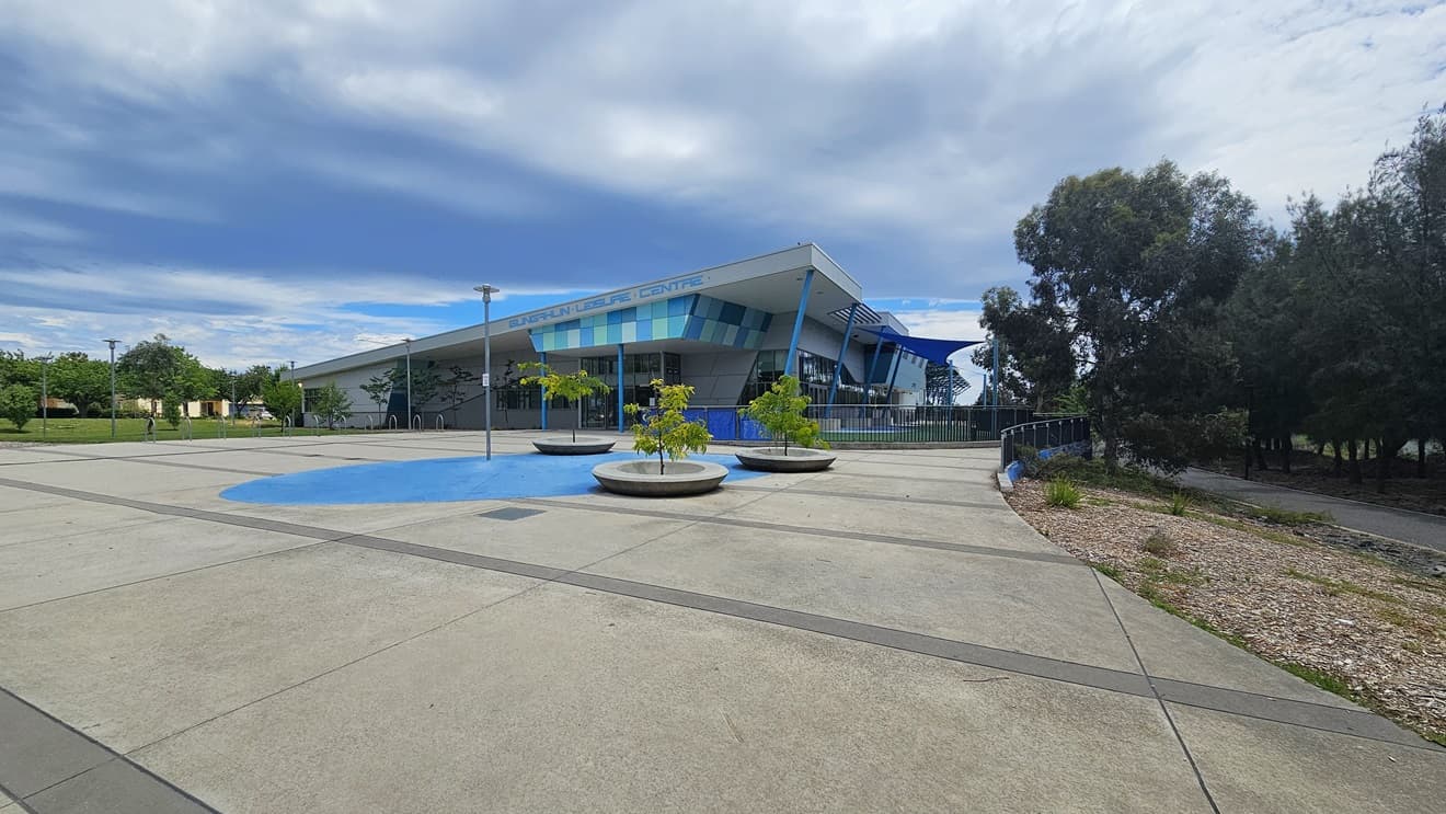 The building is a low-rise concrete structure with contrasting blue trim, and the facility name is displayed at the entrance. The building's concourse features a spacious concrete area. On the left, bike racks are positioned near the grass area. To the right, three small trees are in large concrete pots. The building has a blue fence railing and blue shade cloths. Tall trees and a descending slope fill the area on the right.