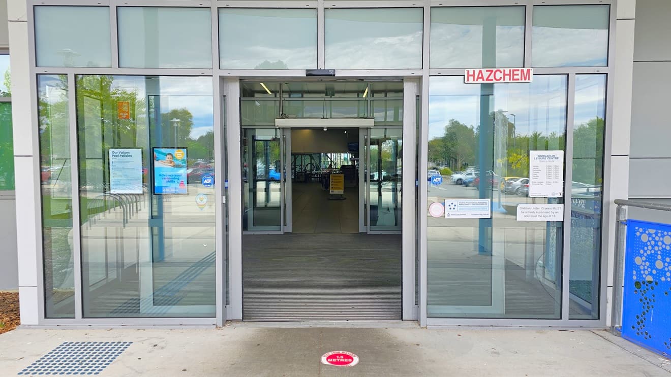 The entrance has double automatic glass doors. Truncated domes are installed on the left. Banners and reminders are affixed to the glass wall. To the right is a blue railing. Inside, a free-standing banner stands in the middle.