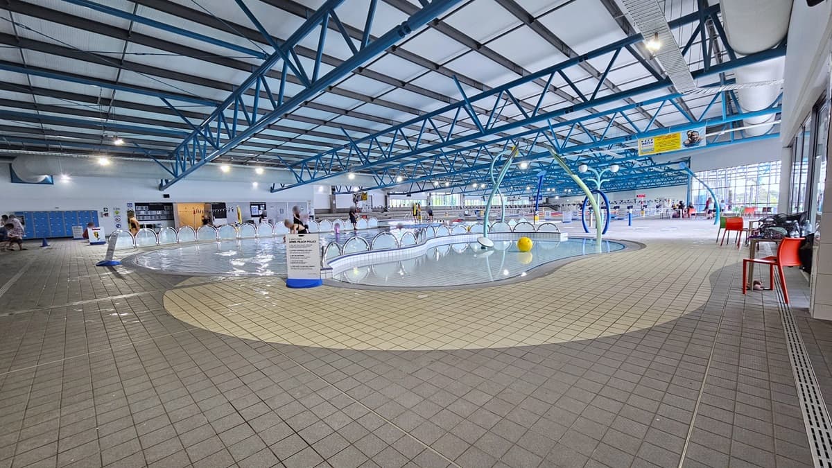 In the middle is the Toddler Splash Pool, equipped with water sprays and fountains. Adjacent to it is the Training Pool, separated by a half-circle fence. On the right, movable plastic chairs and tables are positioned near the glass wall. On the left, blue lockers are against the wall near the accessible change rooms. The ceiling features a white industrial look with exposed blue metal beams. The pool is occupied by guests.