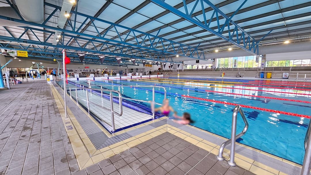 The 50m Lap pool has a ramp entry with handrails and floating ropes. To the far left is the kids' splash pool. Free-standing banners line the pool deck, and tiered seating is available on the far right. Glass walls surround the area, and the ceiling has an industrial look with exposed black and blue metal beams, large aircon vents, and LED lights. Two people are seen on the pool deck. Poles hold a string banner that runs across the pool.