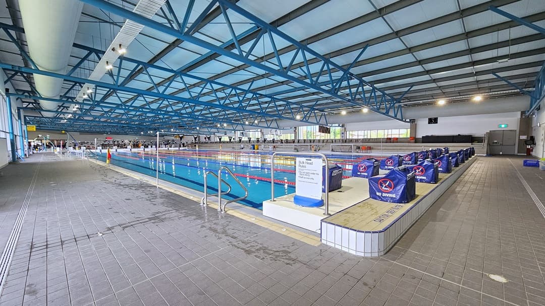 The 50m Lap pool features a ramp entry with handrails and floating ropes. To the right, diving boards with blue covers are arranged on the raised pool deck. A handrail is installed on the corner of the pool. To the far right is tiered seating, and to the far left is the kids' pool. Poles hold a string banner that runs across the pool. Glass walls surround the area, and the ceiling has an industrial look with exposed black and blue metal beams, large aircon vents, and LED lights.