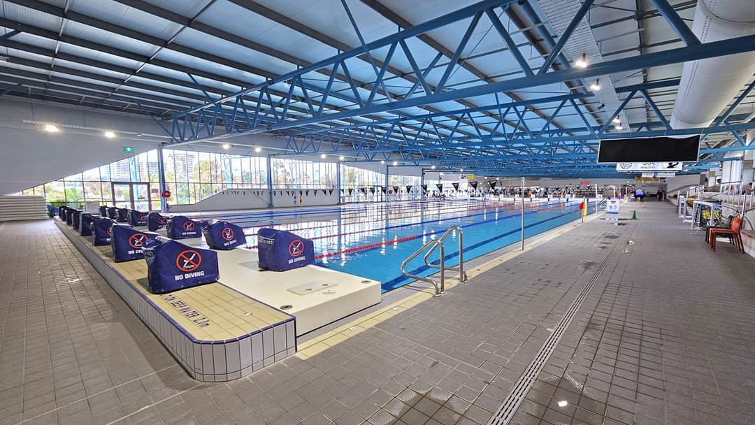 The 50m pool with two sets of dive blocks sitting to the left. Small flags hang over the pool. The blue steel beams are a dominant feature from the ceiling.