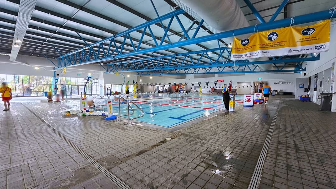 A pool with the dominant blue steel beams hanging from the ceiling. Two lifeguards are walking around the pool. The colourful kids splaspark is in the background.