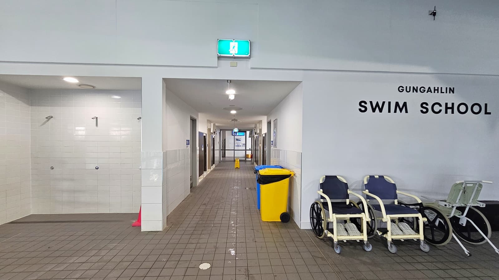 To the left are three showers. Directly ahead is a corridor leading to the bathroom's. A yellow and blue rubbish bin are on the right of the corridor. Three aquatic wheelchairs are placed to the right of the entrance to the corridor. 'Gungahlin Swim School' is written in black on the wall above the wheelchairs.