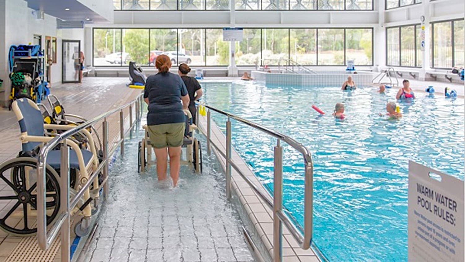 The entrance ramp of the warm water pool has metal handrails to support people with mobility disability while entering the pool. Left of the handrails are two blue and white aquatic wheelchairs. Right of handrails are warm water pool rules to remind the guests of do’s and don’ts.