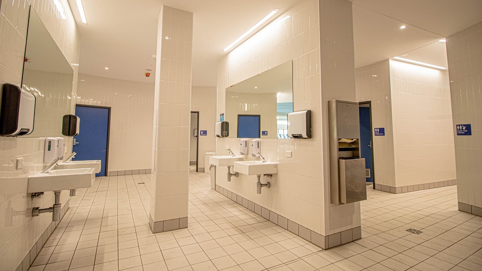 An image showing two separate and clean toilet rooms available at Gurri Wanyara. In this image are 4 washing areas with 4 automatic dispensing soap machines, 2 large mirrors mounted on the walls, and two hand dryers. The toilet doors are painted in blue.