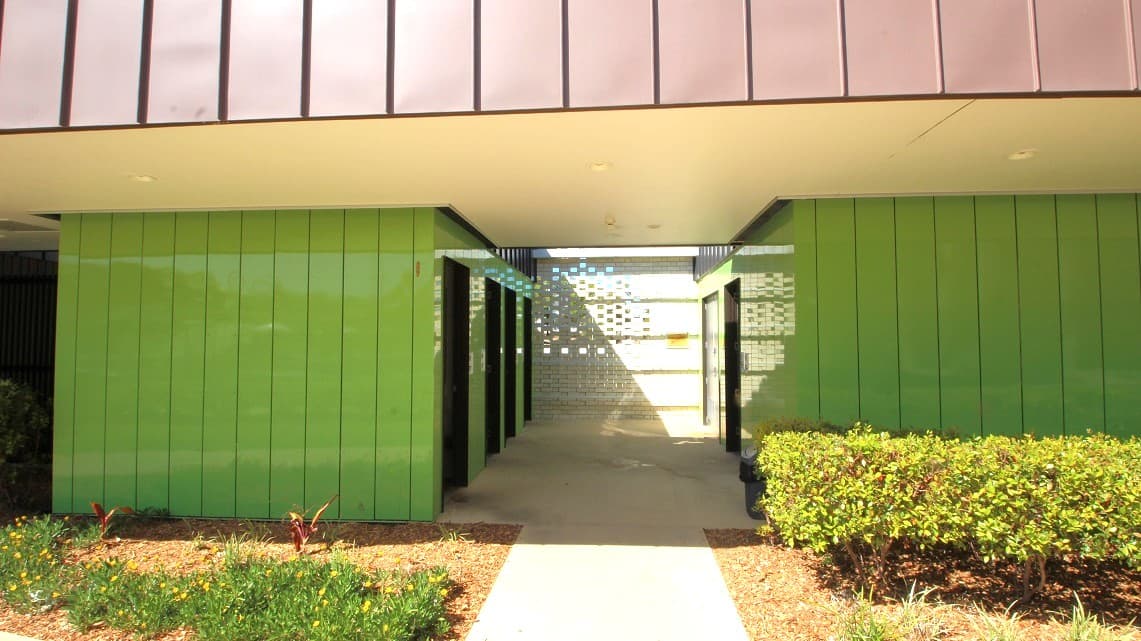 A view of the bathrooms' exterior. There is an aisle between two separated divisions of the lime-green coated painted bathroom walls facing each other mounted with overhead bracings and several standard doors.