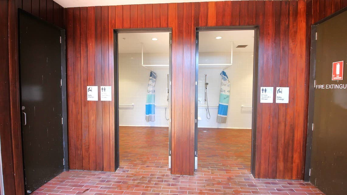 Entrance leading to the bathrooms. Signage can be found on both bathroom doors, conveying that these facilities offer unisex toilets with showers, baby-changing facilities, as well as separate male and female restrooms and showers. Furthermore, there is a rubbish bin located to the right of the restroom entrance. Adjacent to the bathroom, on the right side, there is also a fire extinguisher logo visible.