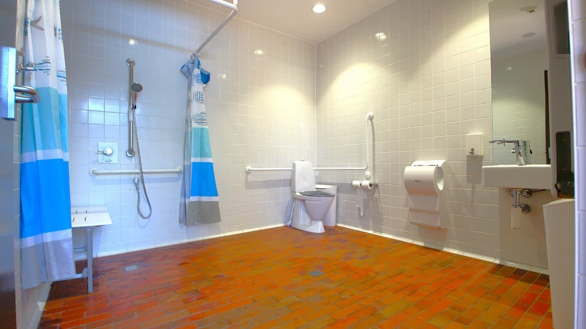 Another view of a bathroom featuring the shower to the left with a blue shaded shower curtain whilst to the right is the toilet. Further to the right is the hand basin and hand dryer.