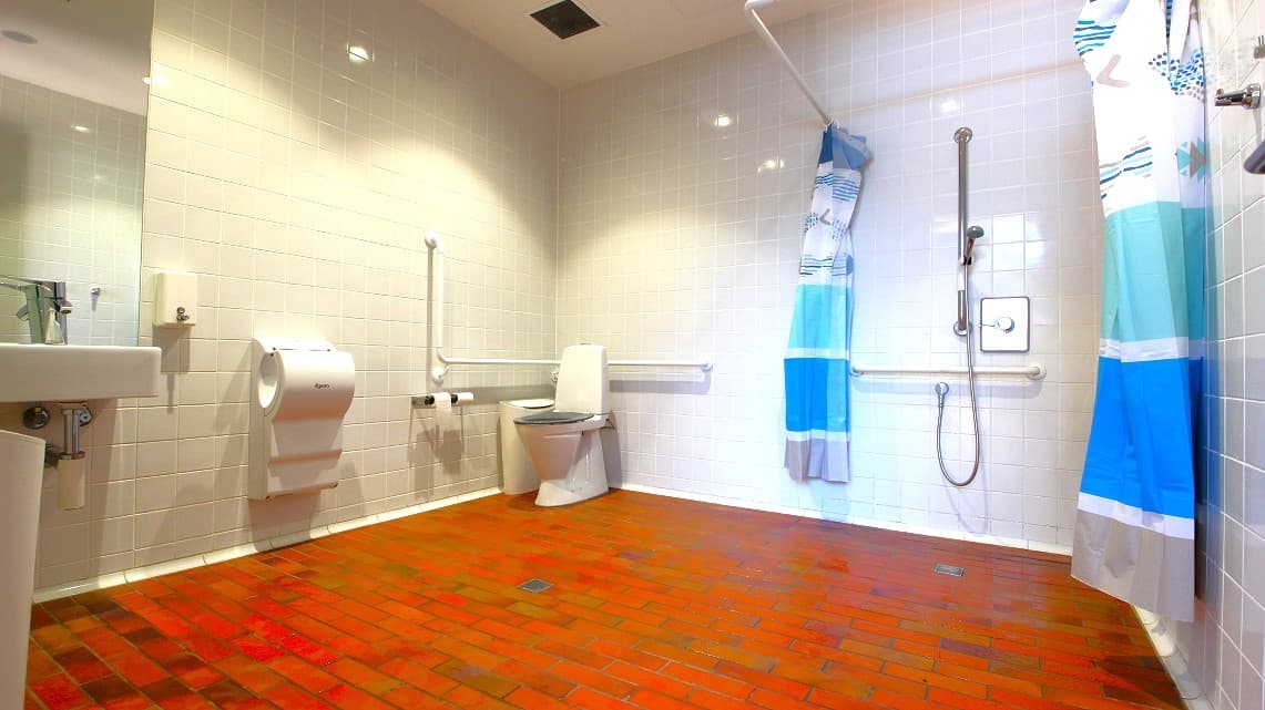 Square ceramic, light-colored tiles cover the bathroom walls. Patterned blue and white curtains draped over the rails served as partitions for privacy. Bright light fills the space overhead through bathroom ceiling lighting. On the other corner is a vanity area of the sink and mirror adjacently positioned to the showerhead and towel racks.