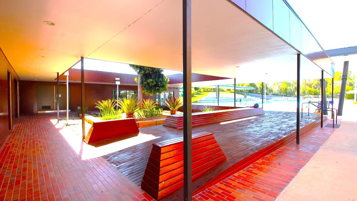 A closer look at the pathways around the veranda. There is seating visible on the spacious open verandas that wrap around a central garden area. Also, this entire section is encompassed by additional gardens. A handrail is clearly positioned in the right corner for safety. Furthermore, fluorescent lighting is prominently installed above the left-side pathway of the veranda, ensuring well-lit conditions.