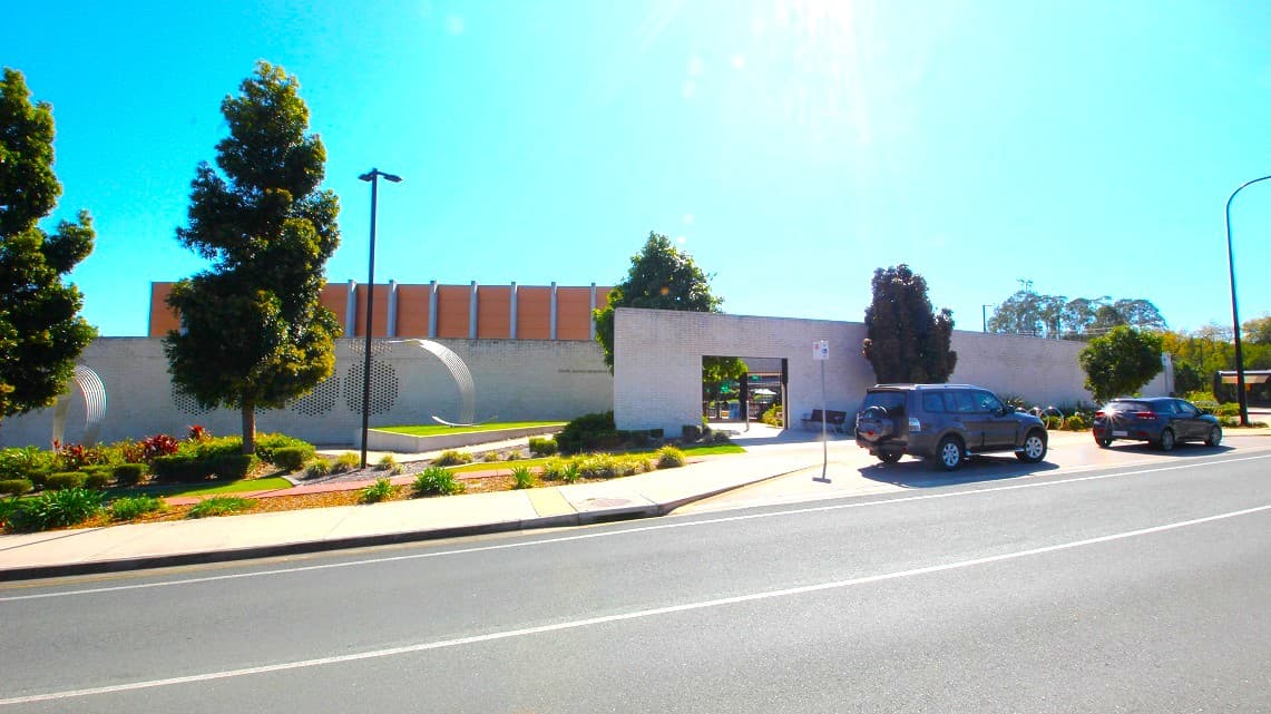 Roadside view of Gympie ARC. The entrance to the Gympie Aquatic Recreation Centre features a distinctive white-brick façade adorned with stainless steel elements. Surrounding gardens and trees further beautify the area, while the color scheme harmoniously blends white, brick, stainless steel, brown, and green elements.