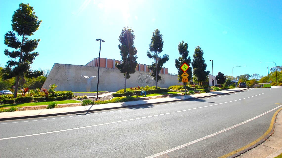 Another angle of the roadside at Gympie ARC. Immaculately manicured gardens and neatly arranged rows of trees grace the surroundings. Alongside these green features, numerous post lights provide illumination, casting a warm glow. Additionally, road signs are conspicuously positioned along the sidewalk, ensuring clear guidance for pedestrians and motorists alike.