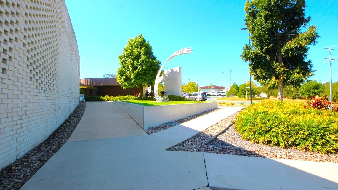 Another angle of the pathways around the Gympie ARC. All three pathways feature identical well-maintained white surfaces, and each one is flanked by neatly placed small rocks, creating a consistent and visually pleasing look. Furthermore, the gardens and trees surrounding the Gympie ARC contribute to the exterior's overall beauty.