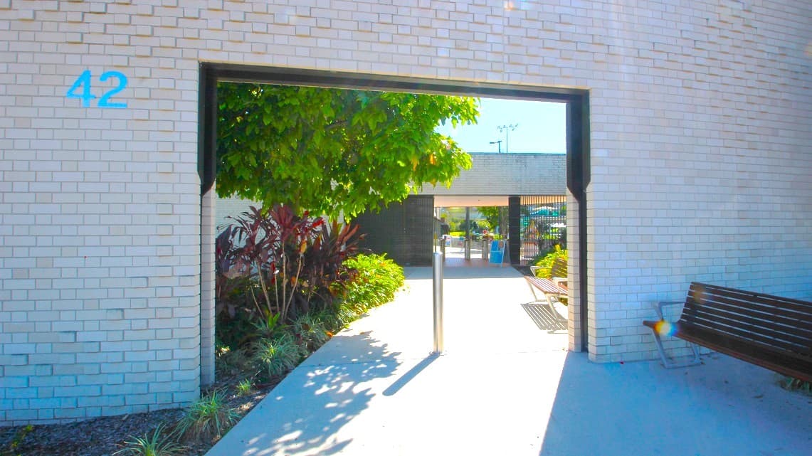 Entrance of the Gympie ARC. As you approach, the white-brick front entrance adorned with stainless steel decorative elements immediately catches your eye, setting a modern and stylish tone. Adjacent to the entrance, there's a metal bench, providing a convenient spot for resting or taking in the surroundings. Notably, the number 42, in a calming blue hue, is affixed to the wall by the front entrance, adding a distinctive touch to the overall design.