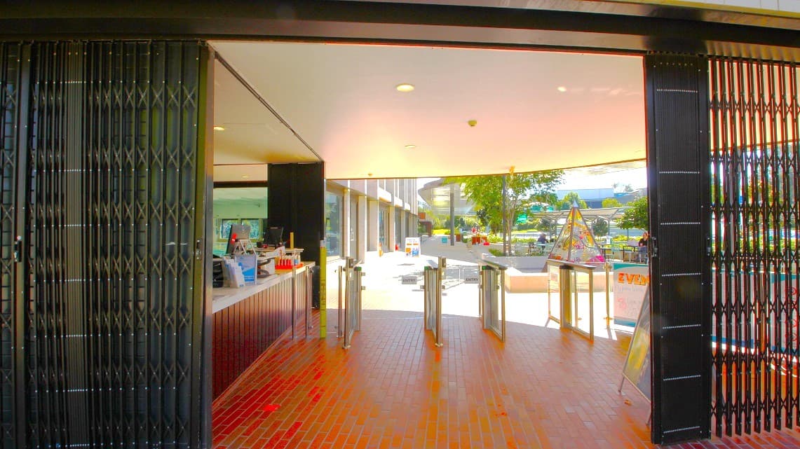 Main entrance of the Gympie ARC. The entrance to the Gympie Aquatic Recreation Centre can be reached through metal grill doors, which are open during the day and securely locked at night. Just to the left of the entrance, you'll find the reception area, a welcoming café, and convenient access to both the indoor pool and the bathrooms. The presence of brick floor tiles adds a touch of durability and aesthetic charm to the space.
