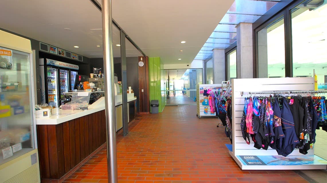 Pathway adjacent to the reception area. To the right, an array of swimwear is elegantly showcased, drawing attention away from the reception area. Inside the reception space, beverages are prominently on display, while framed artwork adorns the walls, adding to the aesthetic appeal and suggesting a focus on both fashion and refreshment.