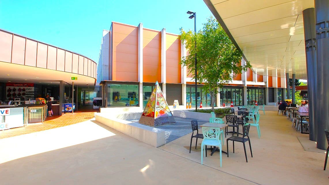 Outdoor area of Gympie Aquatic Recreation Centre. In the center of the area, a quaint small-sized tent stands, providing a focal point. Surrounding it, chairs in various vibrant hues, and tables are strategically placed, creating a cozy seating arrangement. Behind a tree, traffic cones are clearly visible, ensuring safety.