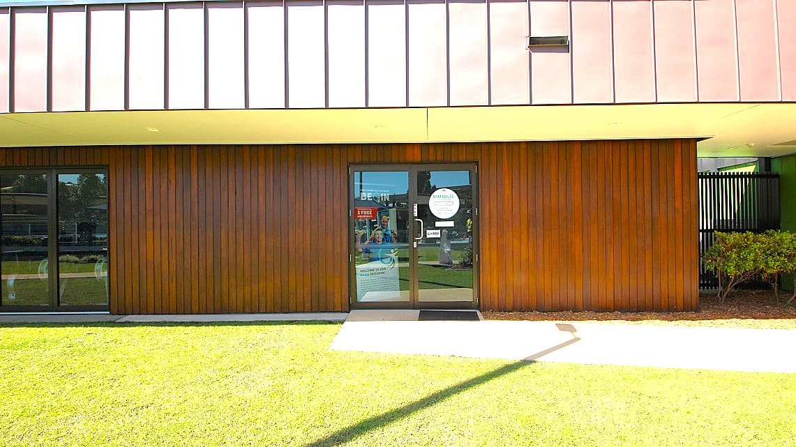 Outdoor look of the Gymnasium’s main entrance. The exterior space is adorned with well-manicured lawns bordering the walkways leading to the gymnasium. The manual glass doors are notably adorned with multiple informative signs. On the right corner, just behind the decorative elements, there stands a tall black fence.