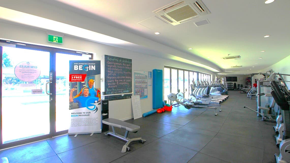 Inside the Gymnasium. The flooring is clad in charcoal-colored rubber matting. Inside the gym, there is a range of fitness equipment, including cardio machines, weight machines, and a dedicated free weight section. The cardio machines are positioned along the left side, running parallel to the windows. On the right side, you'll find additional equipment, like weight machines, arranged in line with the wall's length.