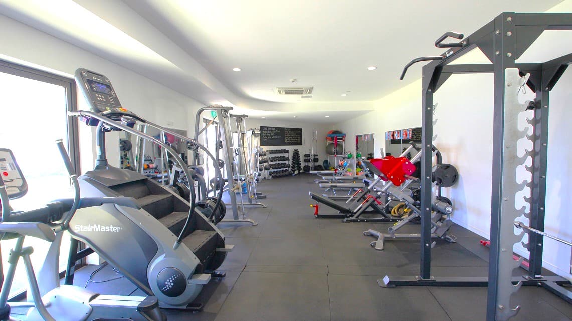 A closer look at the accessible gym equipment. The gym is thoughtfully designed, featuring a well-arranged assortment of cardio machines, weight machines, and a designated space dedicated to free weight exercises. Everything is neatly organized and easily accessible. The walls are painted in a clean and crisp white color, creating a bright and inviting atmosphere. Throughout the entire gym, the illumination is provided by overhead fluorescent lights.