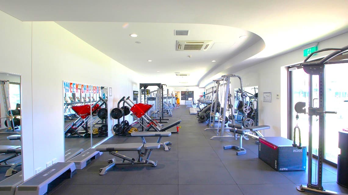 Another angle of the available gym equipment. Various gym equipment, categorized by type, are prominently displayed and neatly organized. Also, the entire floor space is visibly covered by rubber matting. On the lower right side, there's an exit sign positioned above manual glass doors.