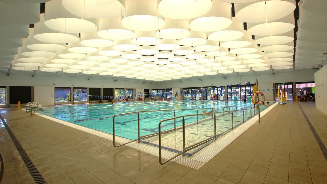 A closer look at the ramp access of the pool. The indoor pool is well-lit, with an array of ceiling lights creating a bright atmosphere. Inside the pool, blue-colored lane ropes and flags are positioned at both ends. These lane ropes separate swim lanes, and the flags help swimmers gauge their position, contributing to a safe and organized swimming environment.