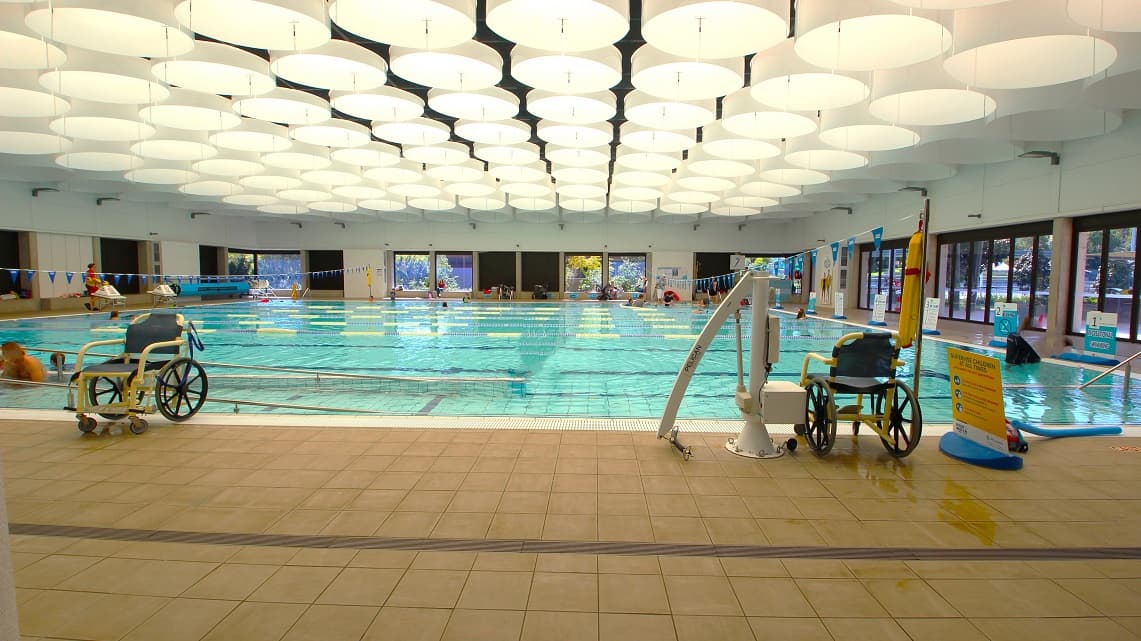 Inside the Indoor Heated Pool. The indoor pool at Gympie ARC spans 25 meters and is entirely enclosed. It maintains a comfortable temperature range between 28°C and 29.5°C and offers access through both ramps and steps. Natural light streams in through glass windows on two sides. Additionally, you can spot a couple of aquatic wheelchairs and a pool hoist, with informative signage positioned next to one of the aquatic wheelchairs.