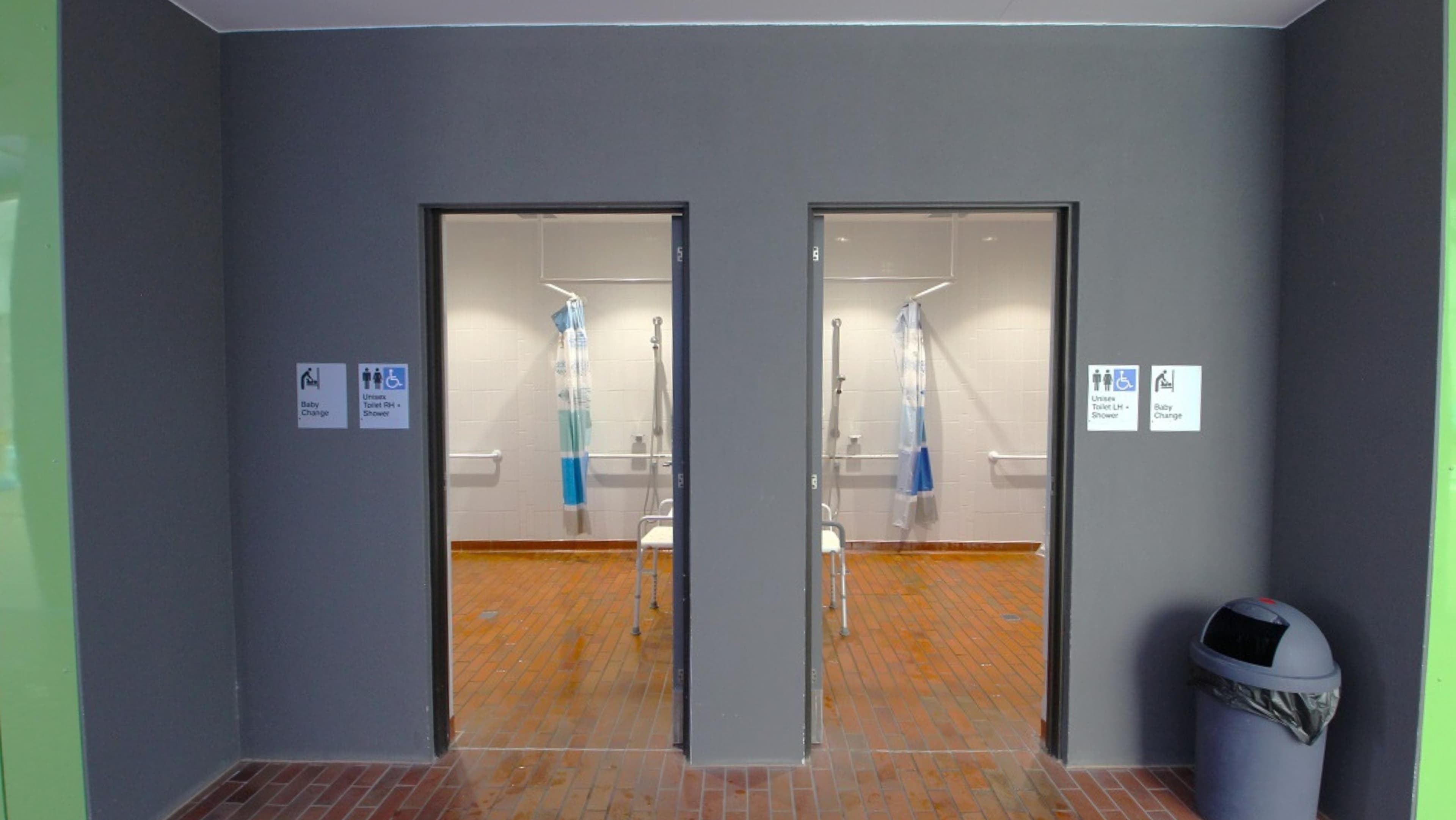 Entrance leading to the accessible bathrooms. Signage is present on both restroom doors, indicating that these facilities provide unisex toilets equipped with showers, baby changing amenities, and male and female restrooms and showers. Additionally, there's a rubbish bin positioned to the right of the restroom entrance.