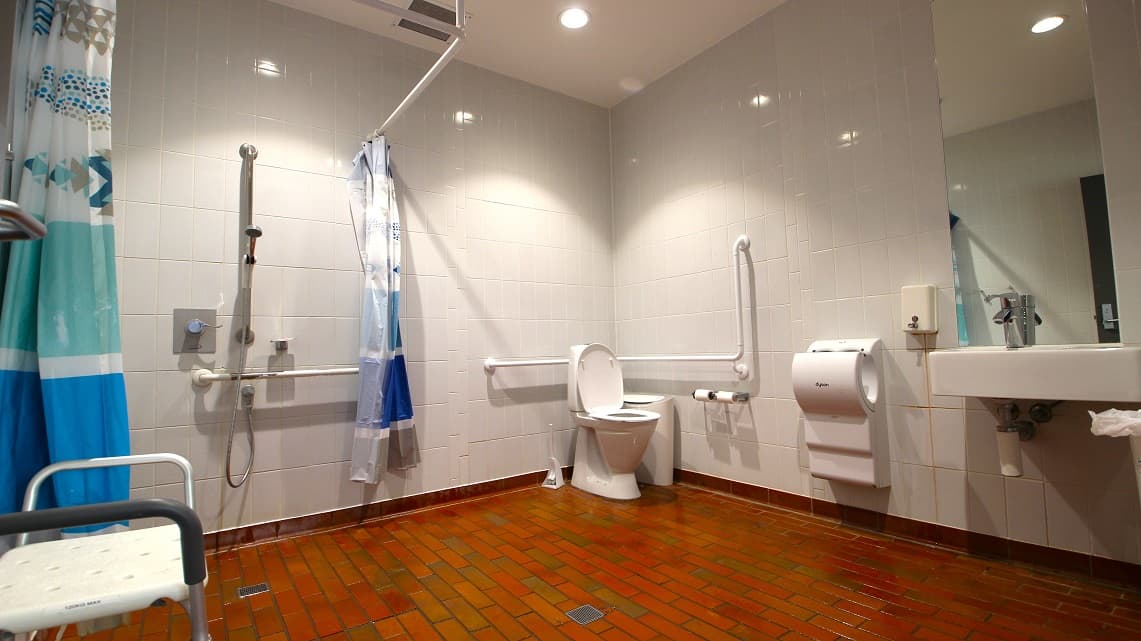 Inside the unisex bathroom. The bathroom distinguishes itself through the use of unique brick floor tiles, making it noticeably different from the surrounding area. Additionally, the toilet, sink, showerhead, toilet paper holder, and towel racks are all placed in a well-organized and spacious manner. Moreover, the bathroom is brightly illuminated with fluorescent lighting, ensuring excellent visibility.