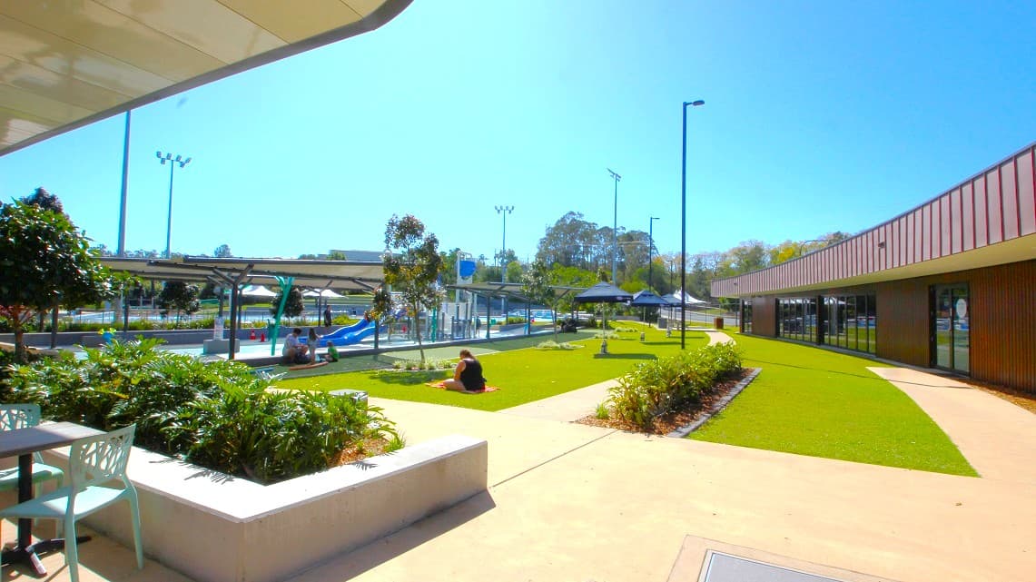 On the left, there is a seating area with a table and chairs next to a planter filled with greenery. In the centre, a person is sitting on a grassy area, surrounded by small trees and plants. To the right, a modern building with large windows runs along the edge of the scene. In the background, there is a playground with slides and a covered area, along with tall light poles and more trees. The sky is clear and blue.