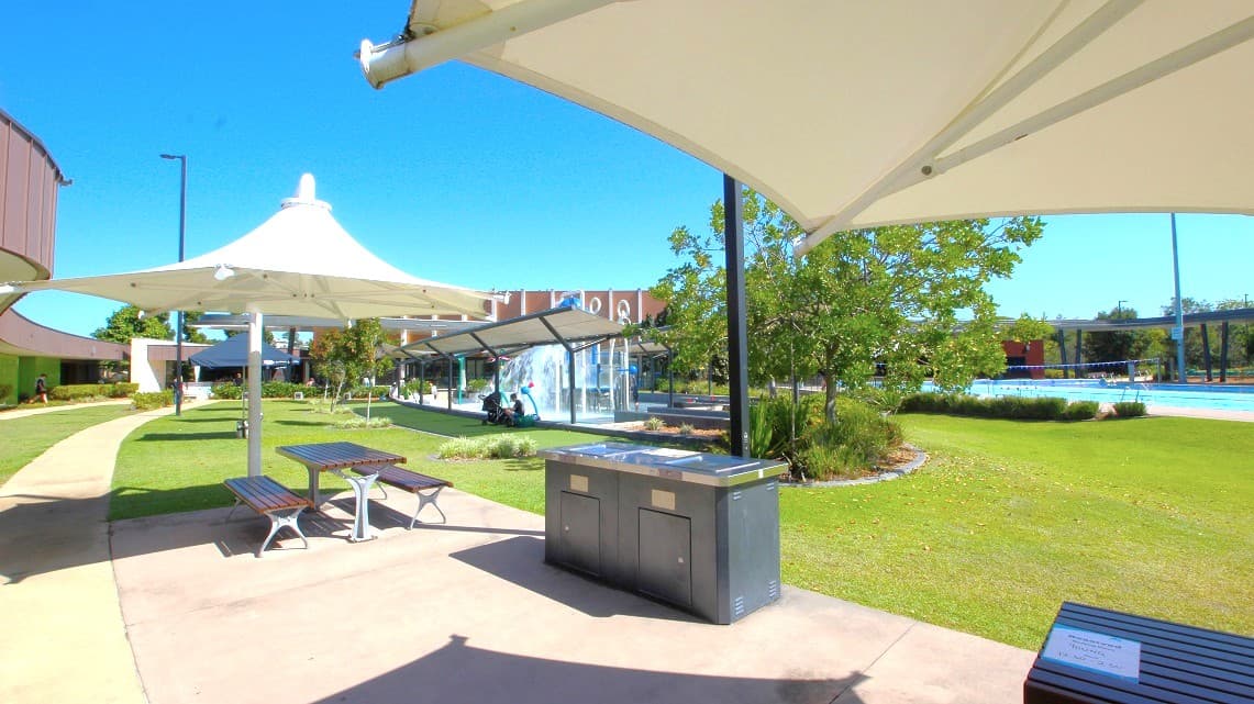 A park area with a paved walkway and green grass. On the left, there are two picnic tables under large white shade structures. In the centre, a barbecue grill is visible. To the right, there are trees and a glimpse of a swimming pool. In the background, a building with a glass facade and more greenery can be seen. The sky is clear and blue.