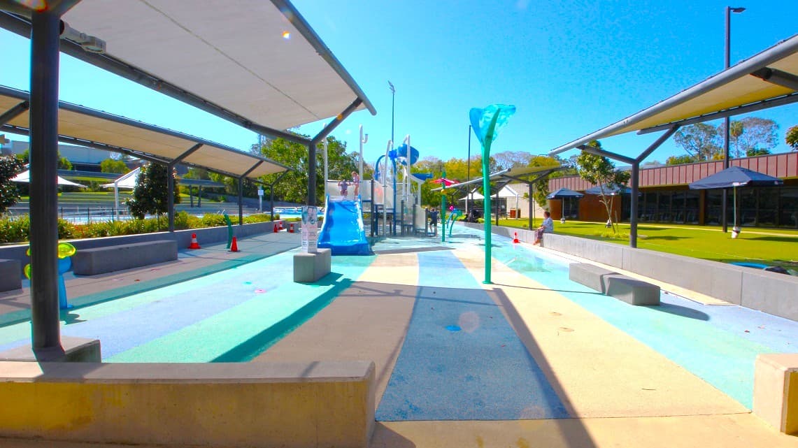 A water play area with a central pathway featuring a blue slide and various water features. On both sides, there are shaded structures with benches underneath. The ground is painted in shades of blue and beige, resembling water. In the background, there are trees and a building with a dark roof. The area is open and sunny, with a clear blue sky above.