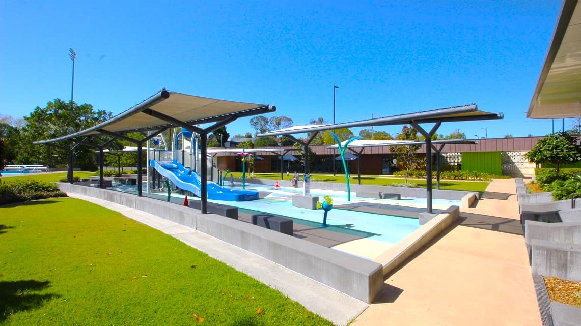A children's water play area with a blue slide in the centre, surrounded by shallow pools. Overhead, there are shade structures with metal frames. The ground is a mix of concrete paths and green grass. In the background, there are trees and a building with a brown roof. A few colourful water play features are scattered around the area.