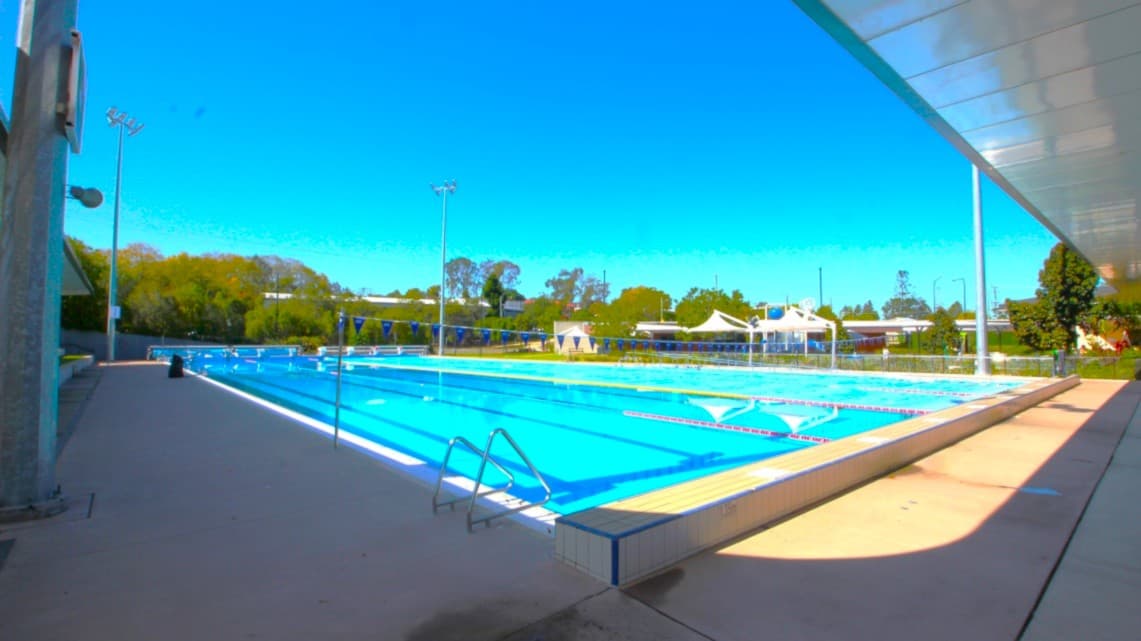 An outdoor swimming pool with clear blue water, surrounded by a concrete deck. On the left, there are tall floodlights and a small building. The right side features a covered walkway with a metal roof. In the background, there are trees and several white tents or canopies. The pool has lane dividers, and there are metal ladders for entry. The area is well-lit, and the sky is clear.
