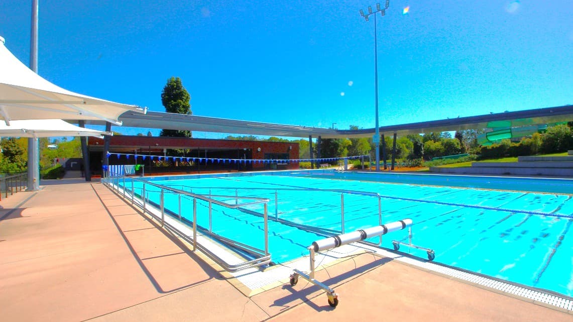 An outdoor swimming pool with clear blue water is surrounded by a paved area. On the left, there is a white canopy providing shade. A metal railing runs along the pool's edge. In the background, there are trees and a low building. A tall light pole is visible on the right. The sky is clear and blue.