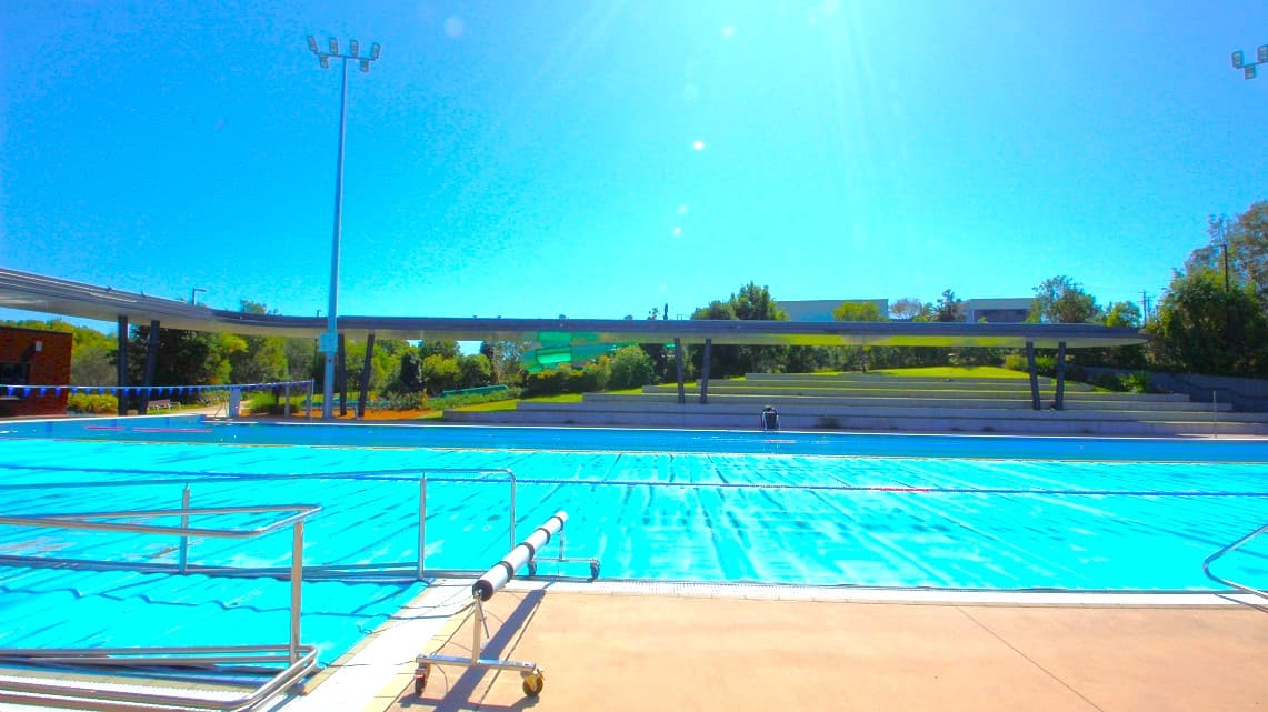 An outdoor swimming pool with clear blue water is in the foreground, surrounded by a beige pool deck. A metal railing runs along the pool's edge. In the background, there is a grassy area with trees and a few structures, including a long, low building. A tall light pole stands near the pool. The sky is clear and bright.