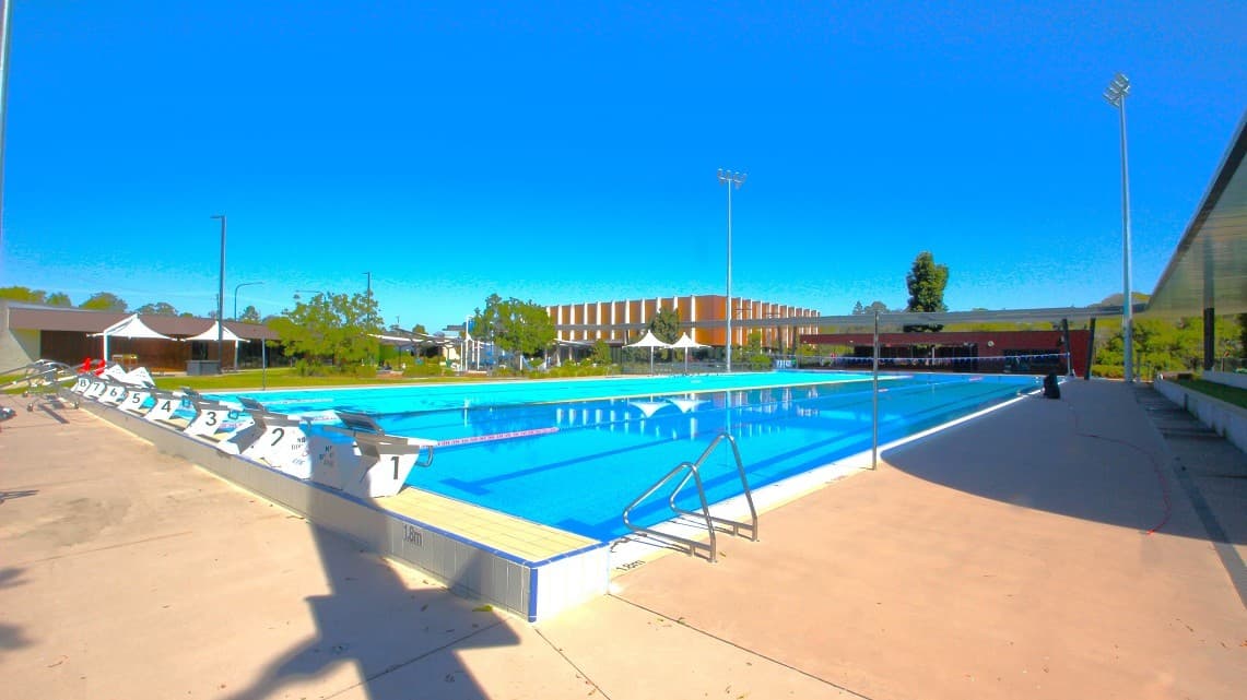 An outdoor swimming pool with clear blue water is surrounded by a concrete deck. On the left, there are numbered starting blocks and a row of white tents. In the background, a large building with a striped facade is visible, along with trees and a few streetlights. The right side features a shaded area with a railing. The sky is clear and blue, and the area is well-lit by tall floodlights.