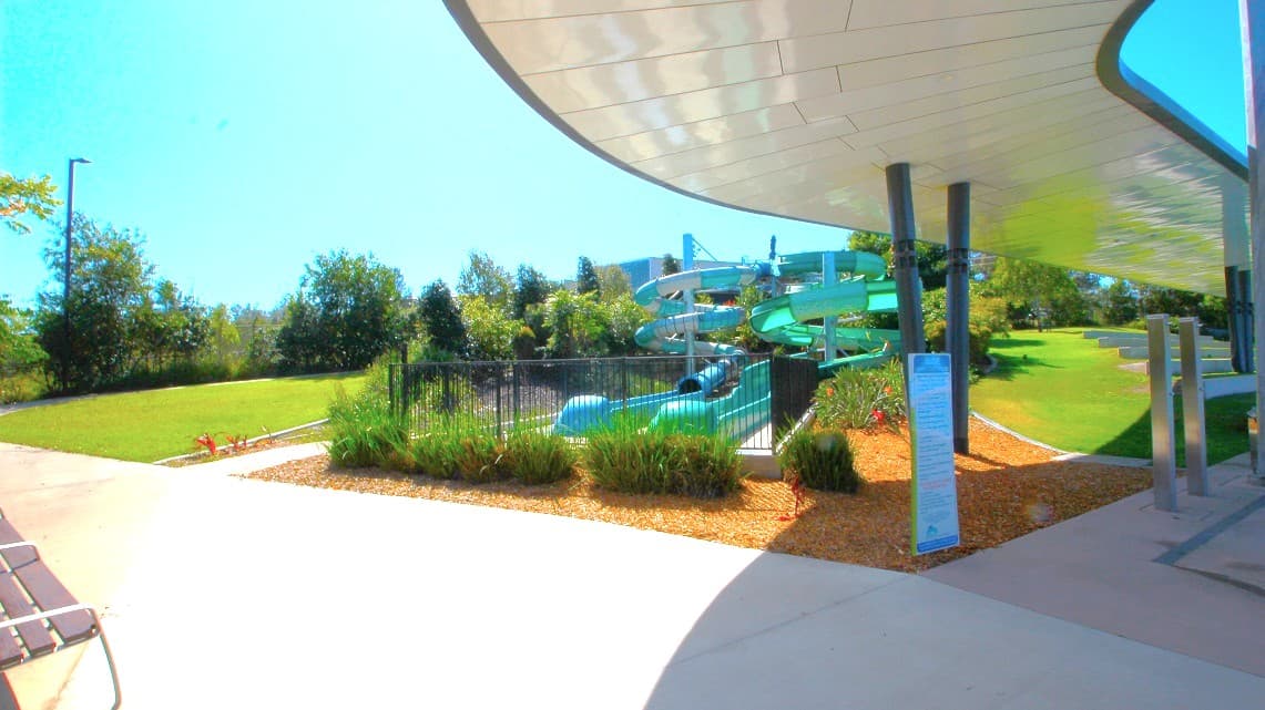 A water park scene with a large, twisting green water slide in the centre, surrounded by a black metal fence and lush greenery. The foreground has a paved walkway and a covered area with a curved roof supported by black pillars. On the left, there's a grassy area with trees in the background. The right side features more grass and a few small structures. The sky is clear and blue.