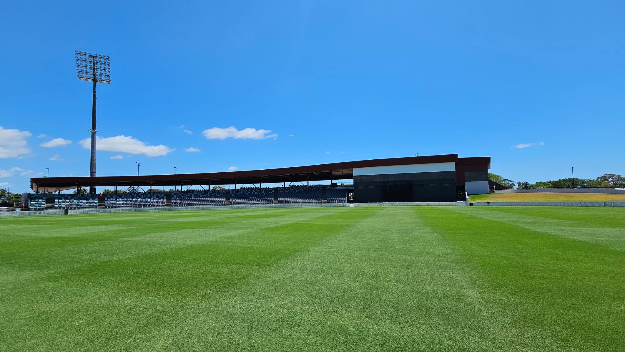 A sports field with well-maintained green grass in the foreground. In the background, there is a long, covered grandstand with rows of seating. To the left, a tall floodlight tower stands prominently. The area around the grandstand is open, with a few trees visible in the distance.