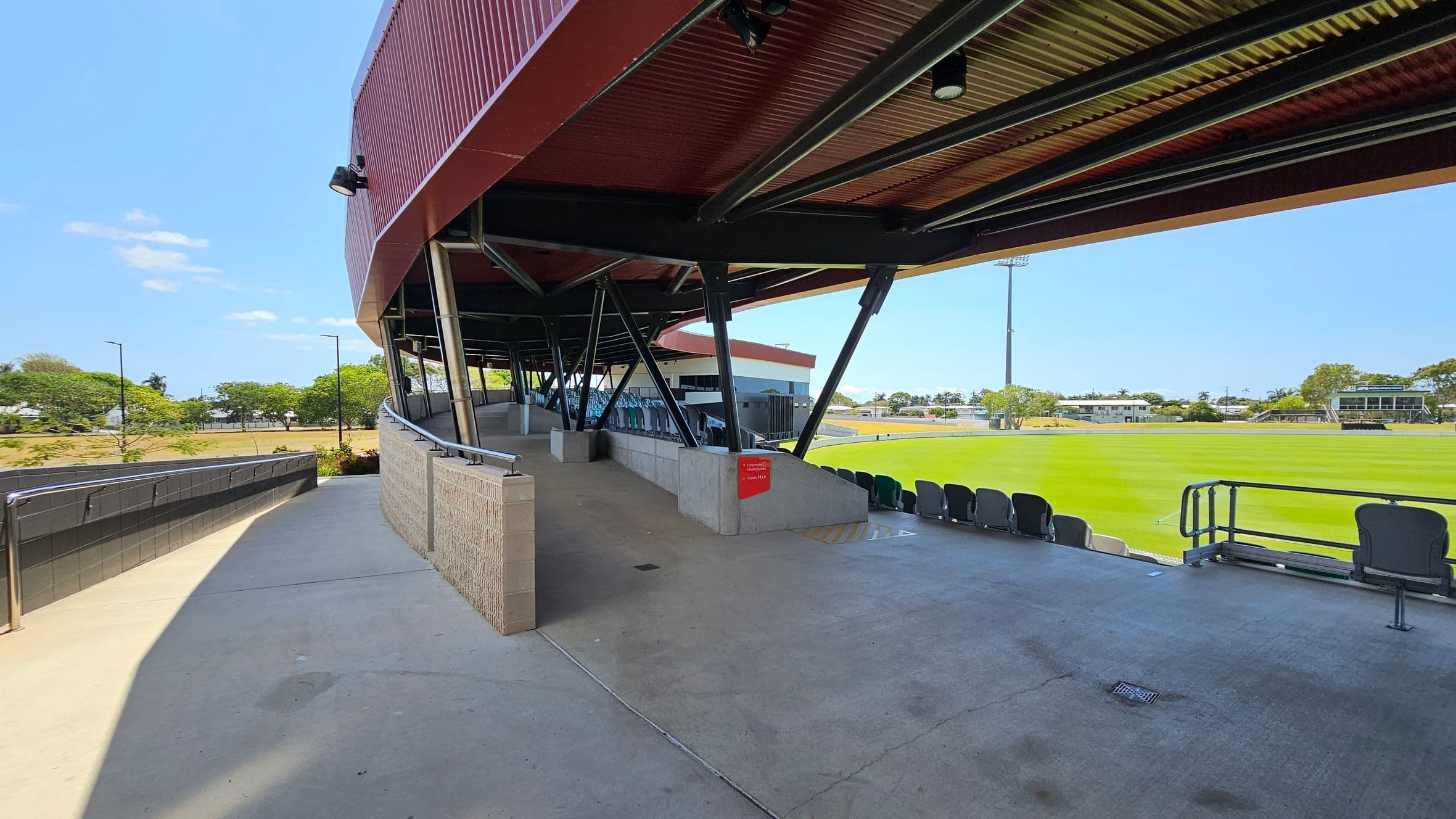 A sports stadium with a covered seating area on the right, supported by black metal beams. The seating area has rows of green and grey seats facing a well-maintained grass field. On the left, a concrete walkway with metal railings leads up to the seating area. In the background, there are trees and a few buildings visible beyond the field. The ceiling of the covered area is made of corrugated metal.