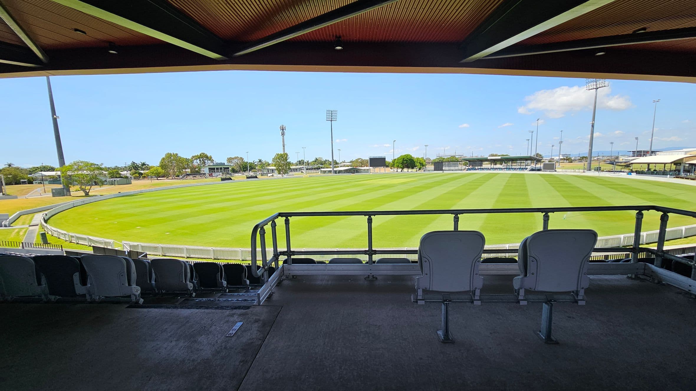 View from a covered stadium seating area overlooking a cricket field. In the foreground, two empty seats face the field. The grass is neatly mowed in alternating light and dark green stripes. Surrounding the field are tall floodlights and a few trees. In the background, there are small buildings and more seating areas. The ceiling above the seating area is wooden with visible beams.