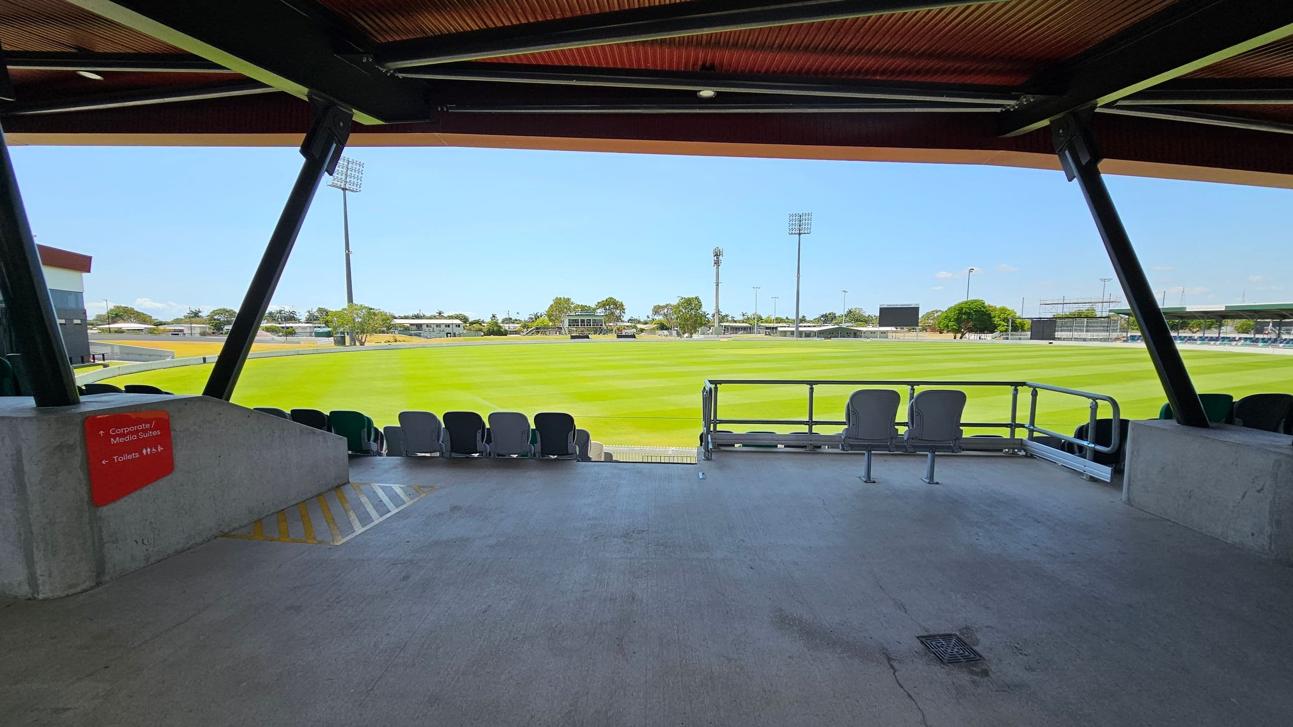 A covered viewing area overlooking a green sports field. In the foreground, there are several empty seats and a concrete floor. A red sign on the left indicates directions to corporate/media suites and toilets. The background features a well-maintained grass field with tall floodlights and trees. The ceiling is supported by black beams.