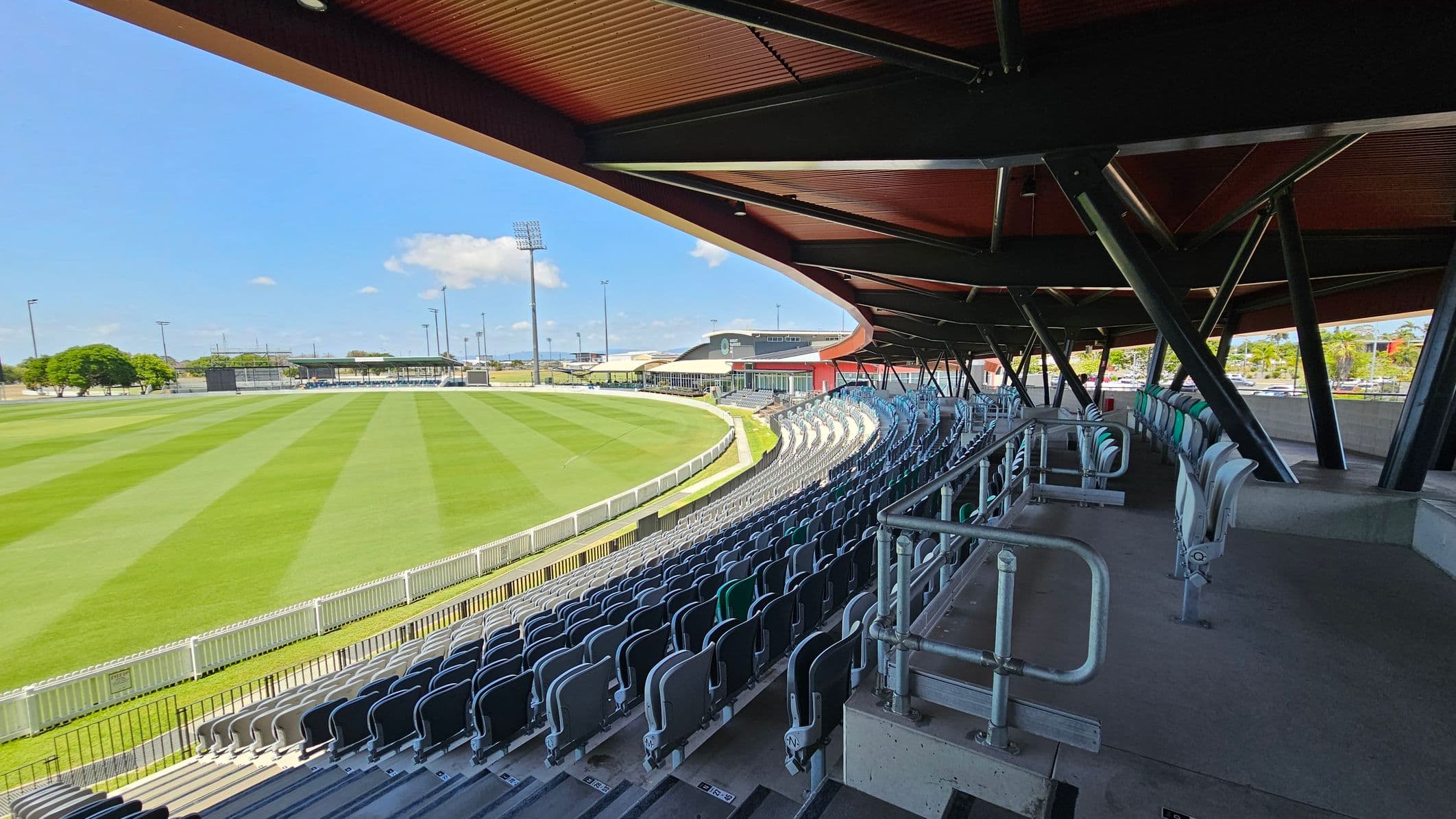 Rows of empty stadium seats curve around the right side, under a large, overhanging roof supported by black beams. The foreground shows metal railings and a concrete floor. On the left, a well-maintained grass field is visible, with white fencing along its perimeter. In the background, there are additional seating areas, trees, and tall floodlights. Buildings and a few parked cars are visible beyond the field.
